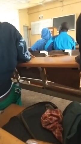 The video shows a classroom setting with wooden desks and chairs. In the foreground, a person wearing a black t-shirt with a white graphic is sitting. Their left arm is raised, and they are wearing a bracelet. In the background, a person wearing a blue hijab and a blue shirt is seated. Next to them, a person wearing a blue shirt is facing away from the camera. A whiteboard is visible on the wall behind them, along with a screen. The lighting appears to be from overhead fluorescent lights. The video is shaky, suggesting it was filmed by someone moving.