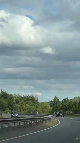 The video captures a view from a vehicle traveling on a highway. The road curves to the right. On the left side of the road, there is a metal guardrail and a dense line of green trees. In the distance, there is a dark-colored car driving in the same direction. Further ahead, to the right side of the road, there is a dark-colored van carrying bicycles on its rear. The van also appears to have a roof rack. The sky is partly cloudy with large, white clouds against a gray backdrop. The overall environment is a highway surrounded by greenery under a cloudy sky.
