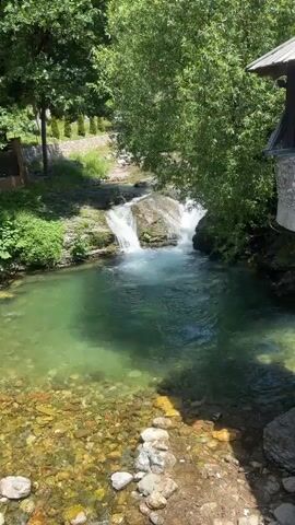 A clear stream flows over rocks with small waterfalls. Lush green trees line the banks of the stream, and the water is a clear green color. The stream bed is visible and filled with stones. A small building with a dark roof is visible on the right side of the frame.