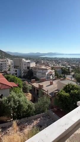 This is a vertical, panning video captured from a high vantage point, likely a balcony or rooftop. The scene overlooks a sprawling coastal city bathed in bright sunlight under a clear blue sky. In the foreground, the edge of a white balcony railing is visible, along with some rubble and construction materials, suggesting ongoing building work. Beyond this, a dense urban landscape unfolds, characterized by numerous buildings of varying sizes and architectural styles, mostly appearing to be residential. Many rooftops are flat, some showing signs of unfinished construction. Lush green trees are interspersed throughout the city, adding natural elements to the urban fabric. Further in the distance, the city extends towards a tranquil body of water, likely a sea or large bay, with distant landmasses and mountains forming a hazy horizon. The sun is prominently visible, shining intensely and casting sharp shadows, indicating it is likely midday. The video pans smoothly from left to right, revealing more of the city and the expansive coastal view. There are no people visible in the video.