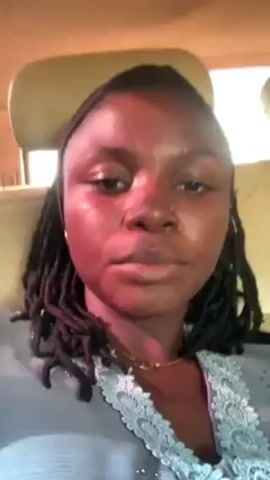 A person with dark skin and braided hair is in the back of a vehicle. The person looks at the camera and touches their chest. The background is blurry, showing the interior of the vehicle including a headrest. The light appears red on the person's face. The person is wearing a gray garment with white lace detailing around the neckline. The vehicle appears to be in motion.