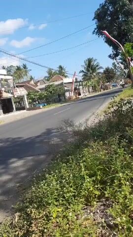 A motorcyclist rides down a paved road. The road passes by several houses on the left side of the frame. Trees and vegetation are visible on the right side of the road, as well as along the left side. The sky is blue with some clouds. A flag with red and white stripes is visible on the right side of the road.