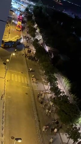 The video shows an aerial view of a street at night, with several yellow buses parked along the side. A group of people are walking along a sidewalk lined with trees and a fence. Some people appear to be sitting or resting on the sidewalk. The street is lit by streetlights, and there is a pedestrian crossing visible. The overall impression is of a gathering or event, possibly a concert or festival, with attendees waiting or departing. The scene is dynamic, with people moving and interacting, suggesting a lively atmosphere.