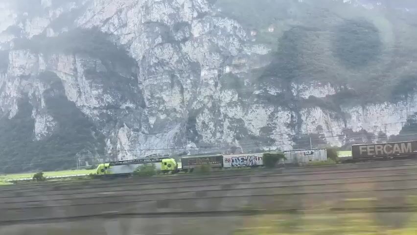 The video shows a train traveling alongside a mountain range. The train appears to be composed of several carriages, including what looks like a locomotive and cargo containers. The mountain landscape is rugged with rocky cliffs and green vegetation. The sky is bright, suggesting daylight, and there are some blurry circles in the footage, possibly reflections or lens flares. The train is moving on a track, and there are also what seem to be vineyards in the foreground, separated by a fence. The scene appears to be rural or mountainous.
