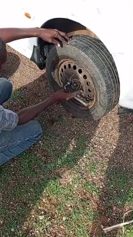 A person wearing jeans and a plaid shirt kneels next to a white car in a grassy area covered in small pebbles. The person is using a lug wrench to remove the lug nuts from the car's front tire. The tire is black and appears to be flat. The background consists of grass, pebbles, and the side of the white car.
