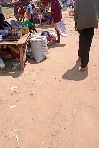 This video shows an outdoor market scene. In the foreground, a person in a red shirt and a pink and white striped bag is standing. To their left, a wooden table displays several bottles of liquid, possibly oil or beverages. Behind the table, a large sack is visible, with text on it that appears to be a product label. More people are seen in the background, some sitting and some standing, near stalls with various goods, including tomatoes and other produce. The ground is dusty and appears to be unpaved. The overall atmosphere suggests a busy marketplace.