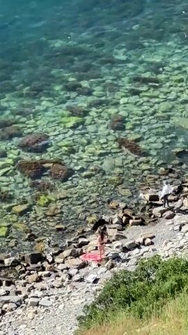 The video shows a rocky coastline with clear, shallow water. Several people are present. On the shore, a person in a bikini is sitting on a pink towel. Further down the beach, another person in a white shirt and dark pants stands near the water. In the water, one person is floating on their back, appearing to relax. The seabed is visible, covered in smooth, varied-colored stones and some seaweed. The water is a turquoise hue.
