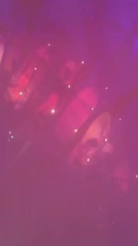 The video shows a low-angle, slightly blurry shot of what appears to be a dimly lit, possibly outdoor, event. The dominant colors are deep pinks and purples, with small, bright white lights scattered throughout the frame, resembling fairy lights or small lamps. In the background, several large, cylindrical objects are visible, possibly lanterns or decorations, with some appearing to have red or pink interiors. The scene is hazy, suggesting the presence of fog or smoke. Several figures, presumably people, are partially visible in the background, some appearing to be sitting or standing. The overall impression is of a festive or atmospheric gathering.