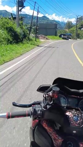 The video is filmed from the perspective of someone riding a motorcycle on a paved road. The road is winding and appears to be in a rural or mountainous area. There are power lines running overhead. On the right side of the road, there is a building under construction, a silver pickup truck, and a blue tarp shelter. The sky is clear and blue with some white clouds. The overall environment appears to be sunny and warm.