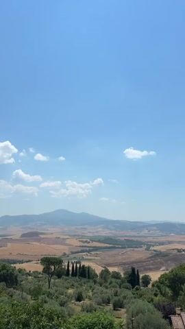 The video shows a landscape view with a clear blue sky and some clouds. The foreground features green trees and bushes, with some cypress trees visible. In the middle ground, there are olive trees and more greenery. The background displays rolling hills with fields of dry grass, suggesting a harvest season. A mountain range is visible in the distance. The overall impression is a rural, possibly Tuscan, landscape on a sunny day.