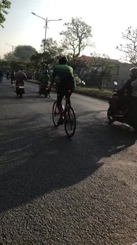 A cyclist in a green shirt and black shorts cycles on a paved road. They are in the center of the frame, moving away from the camera. Behind them, several motorcycles are also on the road, as is another cyclist further in the distance. The scene appears to be outdoors during the daytime, with trees lining the road and sunlight casting shadows. The road surface is asphalt. The video is shot from a low angle, potentially from another moving vehicle.