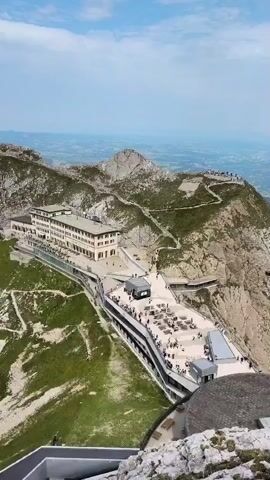 The video shows an aerial view of a mountainous area with buildings and people. The first building is a long, light-colored structure built into the side of a mountain, with many windows and a walkway along its edge. Below this building is a wider, modern-looking structure with an outdoor seating area filled with tables and chairs, and many people are visible sitting and standing there. Further up the mountain, on a rocky peak, a group of people are gathered, and a cross is visible. Paths and walkways are visible winding up the mountainside, with some people walking on them. The surrounding landscape is mountainous with green vegetation on the slopes, and in the distance, a hazy view of plains or valleys with scattered buildings can be seen under a bright, partly cloudy sky.