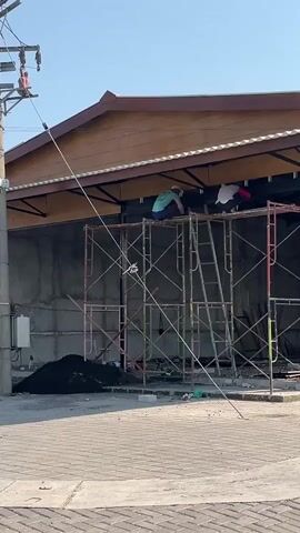 The video shows two workers on scaffolding near a building under construction. The building has a brown roof and a concrete exterior wall. One worker is wearing a blue shirt and the other is wearing a white shirt with red accents. They appear to be working on the exterior of the building, possibly installing a window or facade. There is a pile of black material on the ground near the scaffolding, possibly sand or gravel. A power pole with wires runs diagonally across the frame in front of the building. The sky is clear and blue.