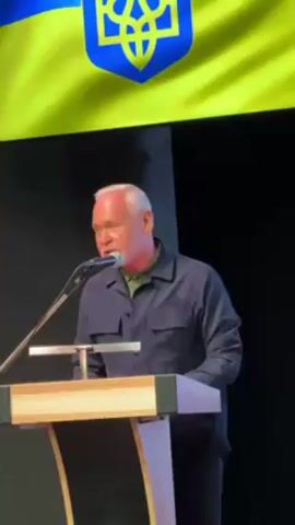 A medium shot captures a white-haired man behind a light-colored podium with dark accents, set against a backdrop of the Ukrainian flag and a black wall. He is wearing a dark blue collared shirt and appears to be reading from notes on the podium, speaking into a microphone. The lighting is bright and focuses on the speaker and the podium.