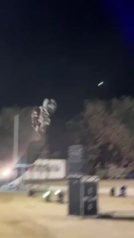 The video shows a motocross stunt at night. A rider is performing a backflip on a motorcycle in the air, illuminated by a green spotlight. The background is dark, with trees and buildings visible in the distance. Another shot shows the rider approaching a ramp on a motorcycle, with a crowd of spectators gathered behind a barrier. A sound system with speakers and lights is also visible in the foreground. The scene appears to be an outdoor event at night, possibly a festival or competition.