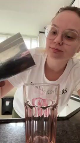 A woman with blonde hair and glasses is pouring a pink smoothie from a metal shaker into a tall, pink-tinted glass on a black countertop. She is wearing a white t-shirt. A trash can is visible in the background to the left, and a tiled backsplash is visible behind the countertop. The shaker is silver and appears to be stainless steel. The glass has vertical ridges and pink details around the rim and the base.