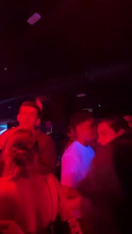 The video shows a dimly lit nightclub or bar. Red and blue lights illuminate the scene, casting a moody atmosphere. Several people are visible, some standing and talking, others potentially dancing. In the foreground, a woman with dark hair is seen from behind. To her right, a man in a light-colored shirt is partially visible. Further back, more people are scattered throughout the venue, with disco balls hanging from the ceiling. The environment appears to be a social gathering space with a bar area and possibly a dance floor.