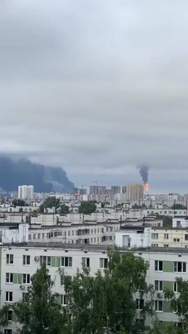 The video shows a cityscape under a cloudy sky. In the distance, large plumes of dark smoke rise from what appears to be a fire, with a visible flame jetting upwards from one location. The smoke drifts across the horizon, partially obscuring taller buildings. In the foreground, there are multiple residential buildings, some appearing to be apartment blocks with balconies and air conditioning units. Trees with green foliage are visible in front of some of the lower buildings. The overall impression is one of a developing emergency situation impacting an urban area.