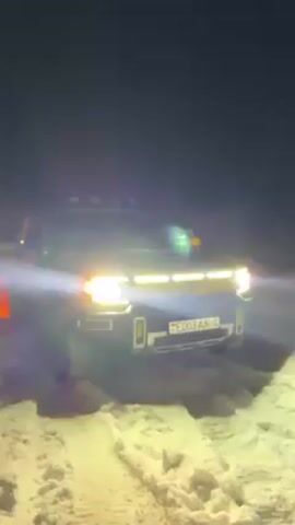 In a night scene, a dark-colored electric pickup truck with bright headlights and roof lights is parked on a snow-covered area. The license plate, visible in the center of the truck’s front, reads “E003 AK-4”. The truck is stationary, and the snow around it appears undisturbed except for the tire tracks. The background is dark, with only faint light sources visible in the distance. The overall lighting is provided by the truck's headlights, which illuminate the snow directly in front of it.