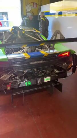 The video shows the rear of a green, blue, and yellow Lamborghini race car with the number 19 on it, inside a garage. Two people are working on the car. A man in a black shirt and sunglasses walks by the car. The garage door is open.