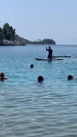A video shows a clear blue bay with several people in the water and one person on a paddleboard. In the foreground, three people are partially submerged in the water, with their heads and shoulders visible. In the middle ground, another person is visible, also partially submerged. In the background, a person is standing on a paddleboard, holding a paddle. The paddleboarder is facing away from the camera. The water is calm and clear, with sunlight reflecting off the surface. The background shows a rocky coastline with green trees and a clear blue sky. The video is a static shot with no movement of the camera.