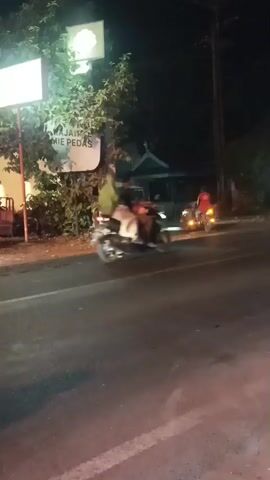 A street scene at night with a food stall on the left. A sign above the stall reads "MAJAPAHIT MIE PEDAS". A person on a motorcycle is seen moving past the stall. Further down the street, a bus is visible. The road is paved and appears to be wet.