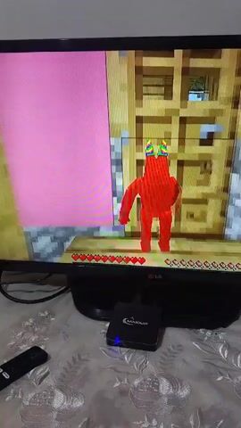 The video shows a close-up of a television screen displaying a video game. The game appears to be Minecraft, with blocky graphics and familiar user interface elements like health bars. In the foreground of the game is a large red character with two black eyes. The character is holding a pink object. The scene in the game transitions to a hallway with yellow and pink walls. Then, the red character is shown standing in front of a wooden door, wearing a colorful hat. The camera then zooms out slightly and the red character is seen from the side, still in front of the door. The video is shot from a slightly high angle, with a tablecloth visible in the foreground and a remote control and a small black electronic device on the table below the TV. The television is on a black stand.