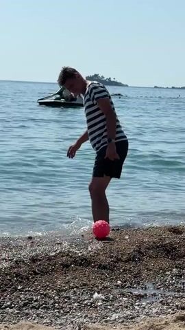 A man on a pebble beach in shallow water is playing with a pink ball. He is wearing a striped t-shirt and black shorts. He kicks the ball, then bends down to pick it up. He then stands up and throws the ball. In the background, there is a jet ski with people on it, and a small island in the distance. The water is blue and calm under a clear sky.