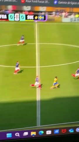 A video shows a soccer match on a laptop screen. A player in a blue and white uniform is dribbling the ball down the field, pursued by a player in a yellow uniform. Other players in blue and white uniforms are also on the field. The scoreboard shows 0-0. In the background, a stadium crowd is visible. The laptop screen is on a desk, with browser tabs visible at the top.