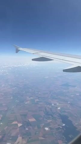 This is a first-person perspective video taken from an airplane window. The camera is pointed downwards and outwards, showing the view of the Earth's surface from high altitude. The airplane's wing is visible in the foreground, extending from the left side of the frame to the right. The wing is white and has a prominent flap or spoiler. Below the wing, the landscape appears as a patchwork of fields, with various colors suggesting different crops or land use. The fields are arranged in geometric shapes, indicative of agricultural activity. Some small clusters of buildings or villages can be seen scattered across the landscape. The Earth's curvature is subtly visible on the horizon, where the land meets the sky. The sky above is a clear, deep blue, with only a few wispy white clouds scattered in the distance. The lighting suggests it is daytime, with the sun illuminating the scene from an unseen angle. The video appears to be a continuous shot with no significant movement other than the subtle sway of the airplane. No people are visible in the video.