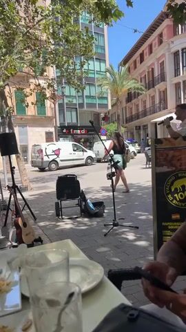 A man is performing music on a sidewalk in front of a cafe. He is holding a microphone and singing. There is a speaker on a stand behind him, and a guitar leaning against another stand to his left. A woman is standing in the background, watching him. There are several white vans and other vehicles on the street behind them. The buildings in the background are tall and modern, with some trees and palm trees visible. A table with plates and glasses is in the foreground, suggesting a cafe setting. The man appears to be interacting with the crowd by holding up an object, possibly a percussion instrument or a prop, while singing.