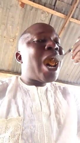 The video shows a close-up, overhead shot of a Black man wearing a white, ornate shirt. He is in the process of eating, with a utensil in his mouth. The background appears to be an outdoor setting, with a corrugated metal roof and wooden beams visible. The lighting is bright, with a strong glare affecting the right side of the frame.