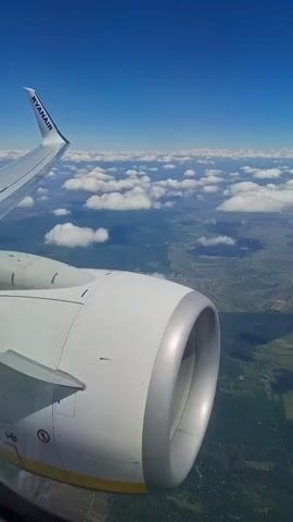 The video shows the exterior view from a Ryanair airplane window. The camera is positioned to capture the aircraft's wing and engine, with the landscape below visible through gaps in the clouds. The sky is a clear blue. The wing, marked with the Ryanair logo, is in the upper-left corner of the frame. The airplane engine is the primary focus, white with a gray intake. The land below is a mix of fields, forests, and small towns, with roads and other infrastructure visible. The presence of clouds partially obscures the landscape. There are no visible people in the video.