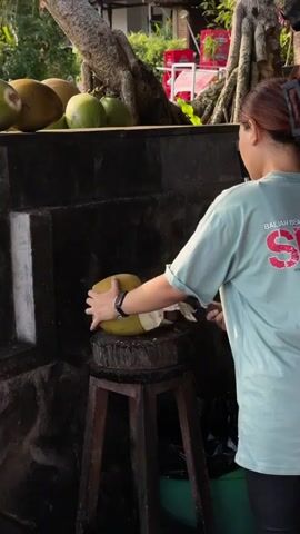 The video shows a person in a light green t-shirt with red writing on the back, holding a large knife and cutting a green coconut. The coconut is resting on a wooden stand. Several other coconuts are visible in the background on a stone surface. The scene appears to be outdoors, with trees and foliage visible. The person is facing away from the camera and is focused on cutting the coconut. The action is captured from a medium distance.