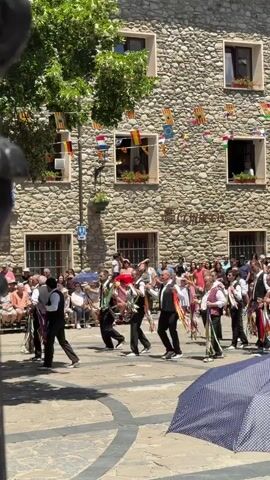 This video captures a vibrant folk dance performance in a town square, likely during a festival. The scene is set in a European-style town with buildings constructed of rough-hewn stone. Small flags, including Catalan and Spanish flags, are strung across the scene, decorating the buildings and suggesting a festive atmosphere. Several windows in the buildings are open, with people looking out, observing the festivities.

The primary subjects are a group of men dressed in traditional attire. They wear white shirts, dark vests, dark trousers, and some have red sashes or neckties. Many of the dancers are adorned with colorful ribbons, which they swing and twirl as they move. They appear to be engaged in a coordinated dance, with many of them playing instruments, possibly bagpipes or similar wind instruments, judging by their postures and the instruments visible. Their movements are lively and energetic, and the overall mood seems joyful and celebratory.

The ground of the square is paved with stone. In the foreground, a large, blue and white polka-dot umbrella is partially visible, suggesting spectators are present and seeking shade from the sun. The time of day appears to be midday, given the bright sunlight and the shadows cast on the ground. The weather seems to be warm and sunny.

The activity taking place is a traditional folk dance and music performance. The men's synchronized movements and the presence of musical instruments indicate a structured cultural event. The surrounding crowd, some seated and others standing, are the audience, participating in the event by their presence and observation. The energy of the dancers and the festive decorations contribute to a strong sense of community and cultural pride.