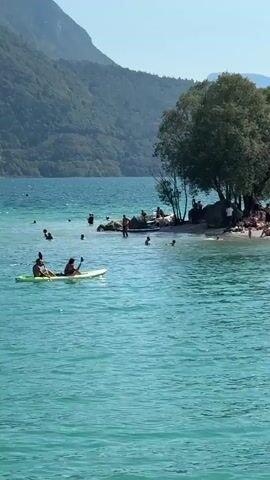 A wide shot captures a bright, sunny day at a lake. The water is a vibrant turquoise, and in the foreground, two people are paddling a green kayak. In the middle ground, a sandy shore is populated with numerous people sunbathing, swimming, and standing. Several trees provide shade on the shore, and a large rock formation is visible. In the background, lush green mountains rise under a clear blue sky. Several other people are swimming or playing in the water.