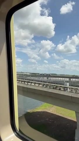 The view from a moving vehicle, probably a train, shows a sunny day with blue sky and puffy white clouds. Below, concrete bridge structures support elevated railway tracks. Below these are railroad tracks. The landscape is mostly flat with some vegetation. The interior of the vehicle is visible in the frame. The view is from inside a window.