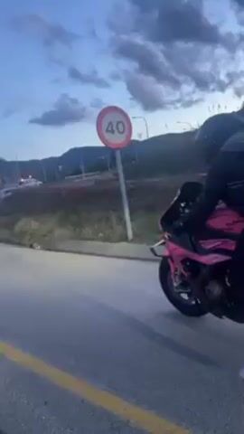 The video shows a person on a pink motorcycle next to a road with a 40 km/h speed limit sign. The person is wearing a black helmet, black jacket, black pants, and white sneakers. They also have a backpack on. The motorcycle has a license plate that reads "68642 LJW". The sky is cloudy and it appears to be dusk.