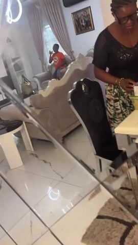 The video shows the interior of a room. A woman in a black and patterned shirt is holding a purple phone, filming. At the beginning of the video, she is standing in front of a mirrored decorative object that reflects a man in an orange shirt sitting on a sofa. There is a picture on the wall behind him. The video then becomes shaky and out of focus as the camera moves around the room. A plant is visible briefly, as well as a black chair. The room appears to be a living room or dining room.