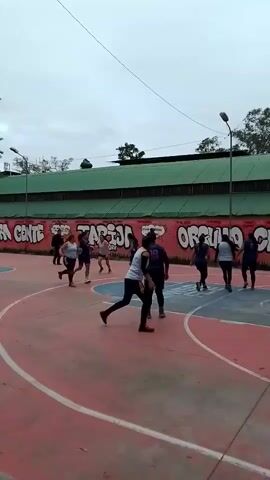 The video shows a group of about ten women playing basketball on an outdoor court. The court has a red and blue surface with painted white lines. The women are dressed in athletic wear. One woman is taking a shot, while the others are positioned around the court. Behind the court is a long, green building with a mural painted on the wall. The mural includes the words "TARIJA ORGULLO CHACO". There are a few trees visible in the background. The sky is overcast.