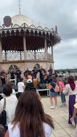 The video shows a band performing on a stage in a gazebo in front of a crowd. The band consists of about ten men playing various instruments, including guitars, drums, and wind instruments. Some audience members are clapping along to the music. One man from the band is dancing on the stage. The gazebo has a distinctive architectural style. The weather appears to be overcast. The crowd is diverse, with people of different ages and styles.