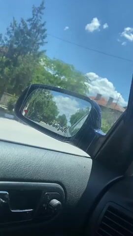 The video captures a first-person view from inside a car, looking towards the side mirror. The side mirror reflects a street scene with buildings, trees, and the sky. A small red heart and the text "love you, drive safe" are visible on the mirror. Another car is briefly visible in the side mirror reflection. The interior of the car, including a door handle and a knob, is in the foreground. The overall setting appears to be a sunny day on a road.