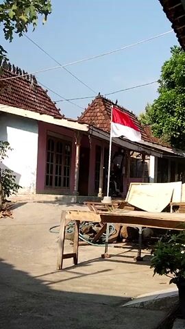 The video shows an outdoor scene in front of a house with a red tiled roof and pink walls. The house has several windows and doors. In front of the house, there is a flagpole with the Indonesian flag (red and white) raised. A person wearing black clothing, possibly a motorcycle helmet, is visible behind the flagpole. In the foreground, there is a wooden structure that appears to be a workbench or table, with various wooden planks and other objects on and around it. There are also trees and foliage visible in the background, and power lines stretch across the sky. The overall environment suggests a rural or suburban setting on a sunny day.