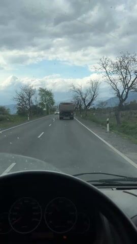 The video captures a journey from a moving vehicle on an overcast day in Pevtsite, Bulgaria.

The initial shot is from the driver's perspective, looking through a smudged windshield at a two-lane asphalt road. A white Mercedes car, with a partially visible license plate "CB 4623", passes on the left. Ahead on the right lane is an older, dark-colored truck. The vehicle's dashboard is prominent in the foreground, featuring a steering wheel and a speedometer displaying numbers up to 170 KM/H. An orange warning light is illuminated on the dashboard. The roadside is lined with bare trees, some with budding leaves, and distant mountains are visible under a cloudy sky.

The perspective briefly shifts to a side view from inside the vehicle, showing a side mirror reflecting the road behind. A yellow lemon and several metal rings are placed on the dashboard. A small red sticker is visible on the interior window pillar.

The final segment of the video shows an exterior view from the right side of the moving vehicle, revealing the road, lush green roadside vegetation, and more trees. A white road marker pole stands beside the asphalt. The sky remains heavily clouded, suggesting an overcast day.