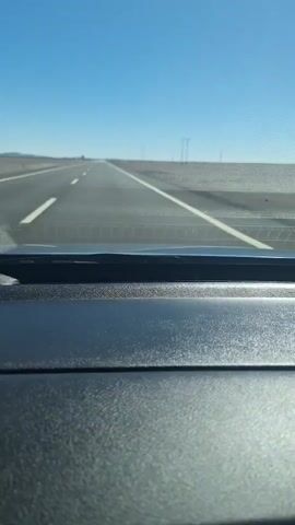 This is a first-person perspective video taken from inside a vehicle, looking through the windshield. The vehicle is traveling on a paved highway with multiple lanes, indicated by white lane markings. The sky is clear and bright blue, suggesting daytime. The landscape on either side of the road appears arid and flat, with sparse vegetation and utility poles visible in the distance. The dashboard of the vehicle is in the foreground, showing a textured black surface. The video captures a sense of continuous travel on an open road.