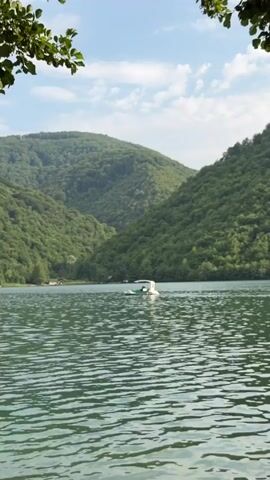 This media file captures a serene view of a lake nestled amidst lush, green, tree-covered mountains. The water's surface is rippled, reflecting the bright, slightly cloudy sky, suggesting a pleasant, perhaps late morning or early afternoon. In the distance, along the lake's edge, small wooden structures, possibly cabins or boathouses, are visible amongst the trees, hinting at a secluded, natural retreat.

The central focus of the scene is a small, white and green pedal boat navigating across the lake. Two people are seated in the boat, appearing to be enjoying a leisurely ride. One individual seems to be steering or pedaling, while the other is seated beside them, possibly taking in the scenery. The overall mood is one of tranquility and peaceful recreation, perfectly complementing the idyllic natural setting. The vibrant green of the mountains and the gentle movement of the water create a sense of calm and escape. There is no visible text in this media file.