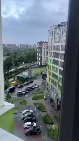 A video shows a view from a window looking out at a city street. The sky is cloudy and it appears to be raining. There are several apartment buildings, a parking lot with cars, and some trees. A soccer field is also visible. The video pans slightly to the left and right.