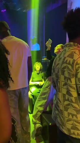 The video shows a nightclub scene with several people visible. A woman in a black hoodie and light-colored sweatpants is holding up a yellow smartphone, presumably taking a photo or video. A woman with curly hair is seated and smiling directly at the camera. In the background, a DJ wearing a white shirt and cap is visible. The lighting in the club is a mix of greens, blues, and yellows, typical of a dance floor ambiance. Other people stand around, partially obscuring the view. A hookah is visible on a table to the right. The atmosphere appears lively and social.