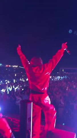 A person wearing a bright red tracksuit stands on a stage with their arms raised, facing away from the camera. They are holding a microphone in their right hand. The stage is lit with bright pink and purple lights, and the background is filled with a large crowd of people, some of whom are holding up their phones with lights on. The sky above is dark, indicating it is nighttime. The person appears to be performing or interacting with the audience.
