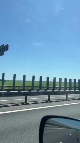 The video captures a view from a vehicle traveling on a highway. On the right side of the road, a series of vertical, grey posts are positioned behind a metal guardrail. Beyond the guardrail, a vast green field stretches out under a clear blue sky. The lower part of the video shows the car's side mirror, reflecting the sky and the road. The sun is visible as a bright light in the sky. The overall scene suggests a journey through a rural or agricultural landscape on a sunny day.